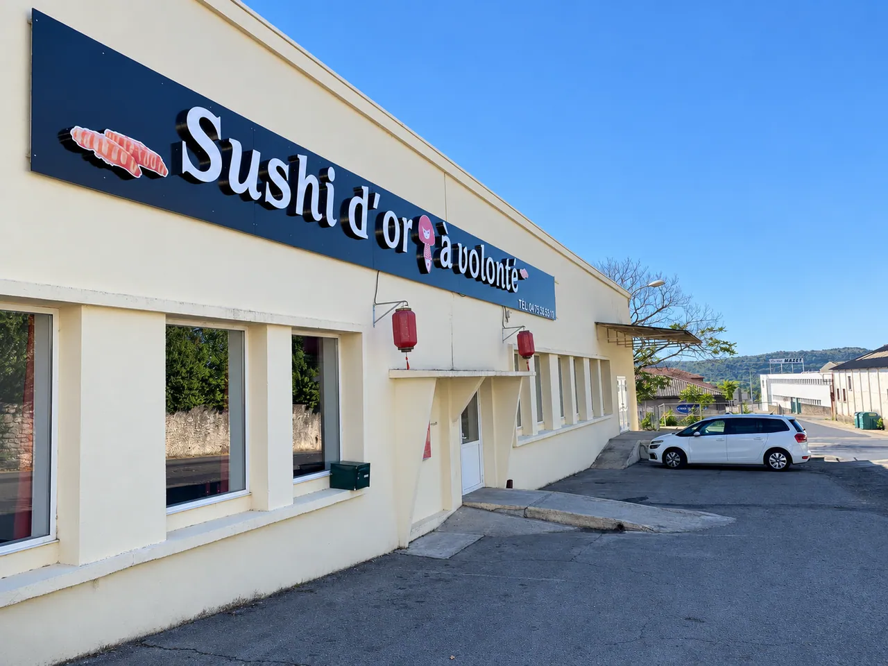 The front of Sushi d'or à volonté: blue sign, red lanterns and car park in front of the entrance.